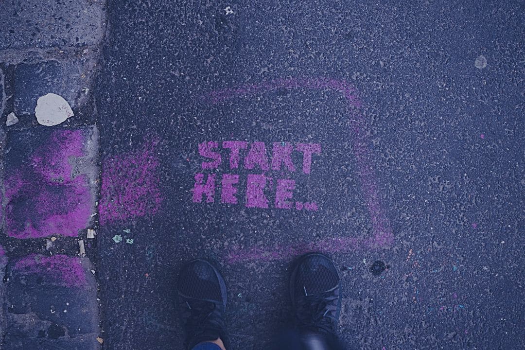 Person standing on street in front of the words "start here" written in pink chalk