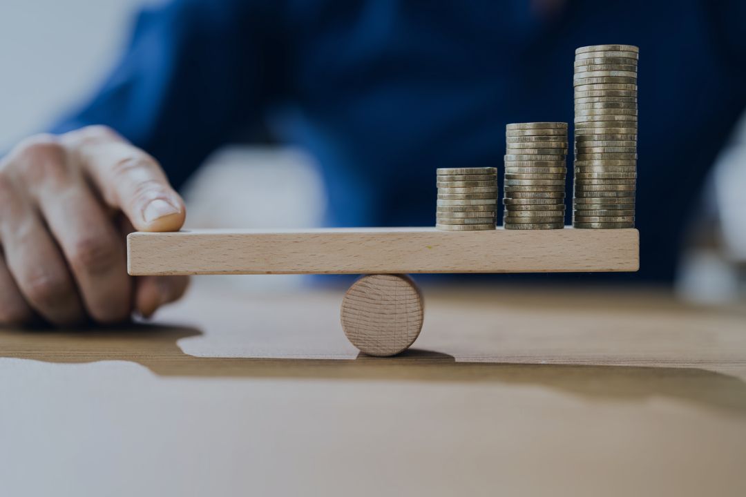 Business man trying to balance toy seesaw with his fingers and stacks of coins